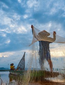 In the early morning before sunrise, an Asian fisherman on a wooden boat casts a net for catching freshwater fish in a natural river.